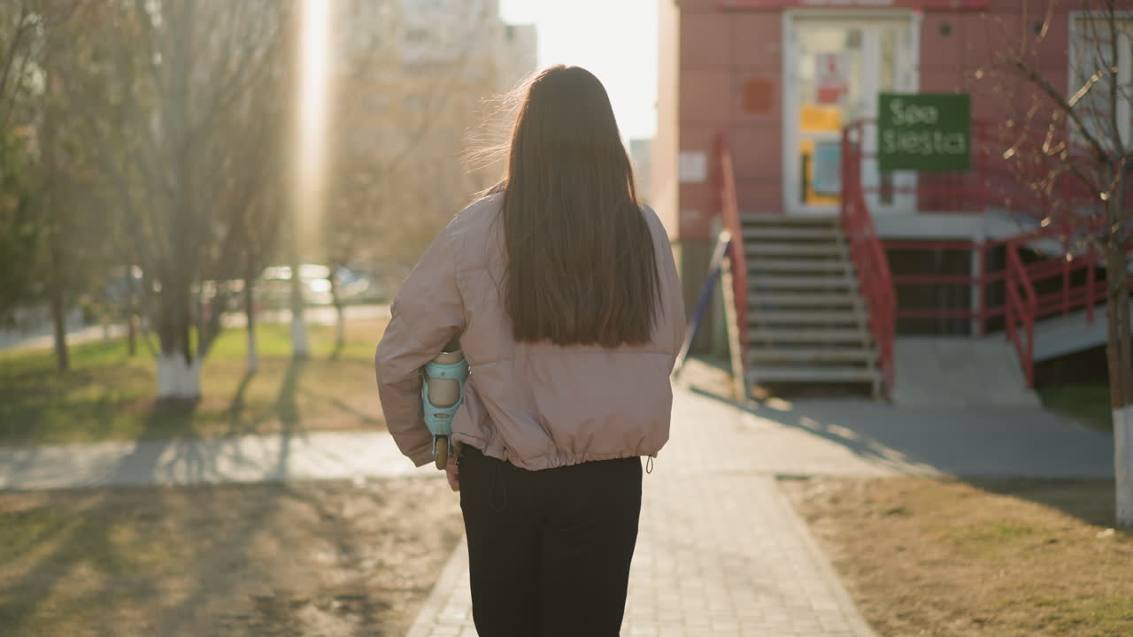 Back shot of a girl wearing a peach jacket and black pants, holding a turquoise skateboard, walking on a sunlit pathway towards a building with sunlight streaming through the trees in the background