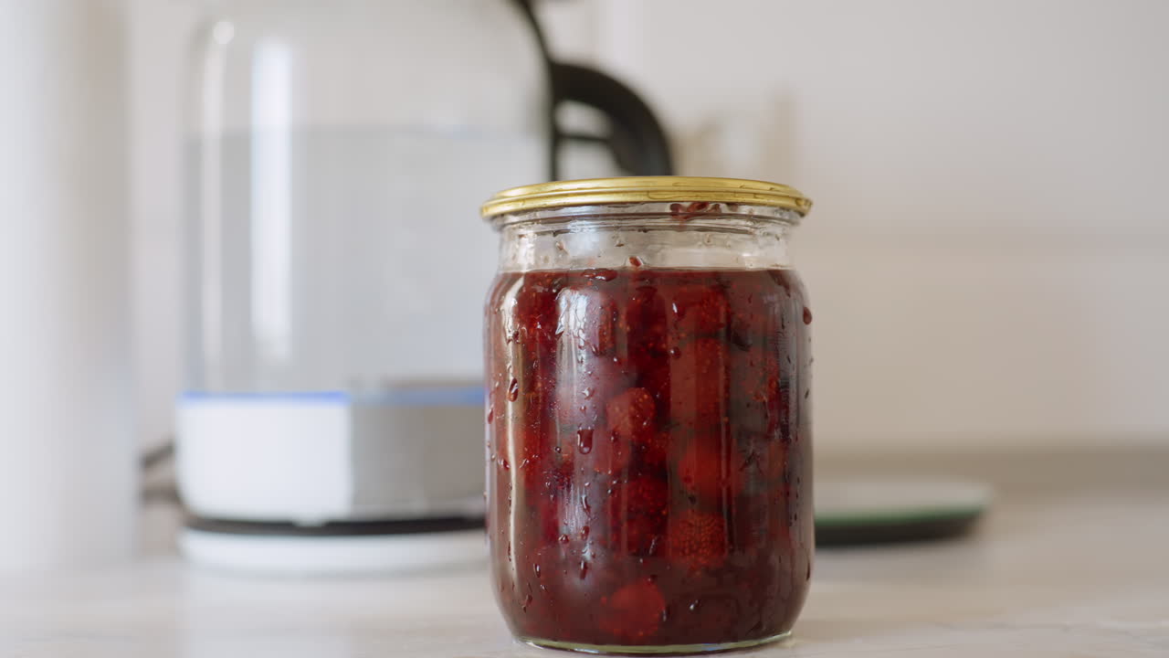Glass jar filled with preserved berries sealed with metal lid stands on clean kitchen counter near electric kettle, with visible condensation and fruit pieces inside