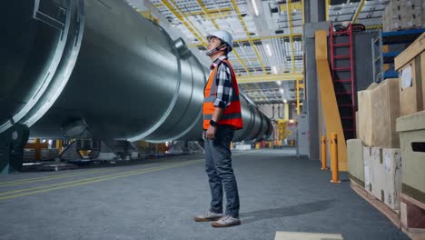 full body side view of asian male engineer with safety helmet standing in pipe manufacturing factory. looking around, checking