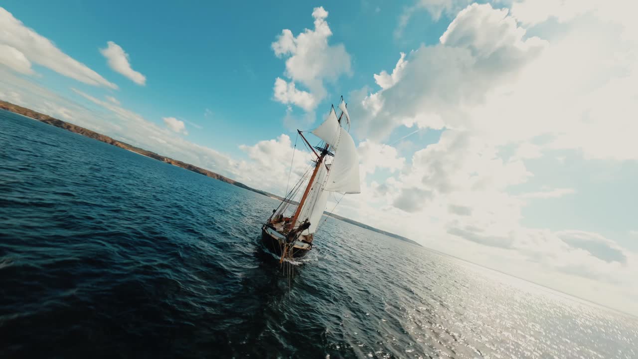 FPV-drone flyver rundt om et tomastet sejlskib, der sejler på blåt hav under solrig himmel.