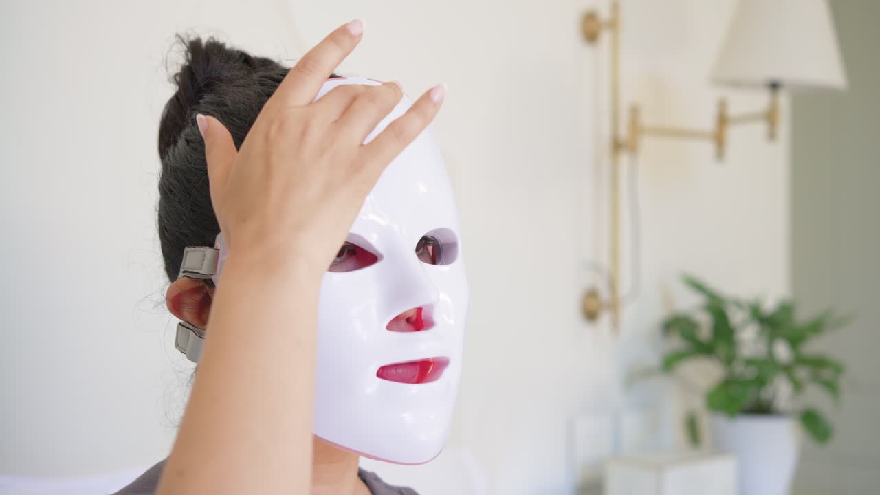 Frau hebt im Schlafzimmer eine weiße LED-Maske zum Gesicht, zieht die Riemen fest, LEDs leuchten zyklisch zur Behandlung.