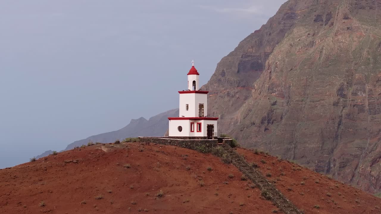 дрон поднимается, захватывая белую часовню на вершине красного холма, прибрежные скалы позади