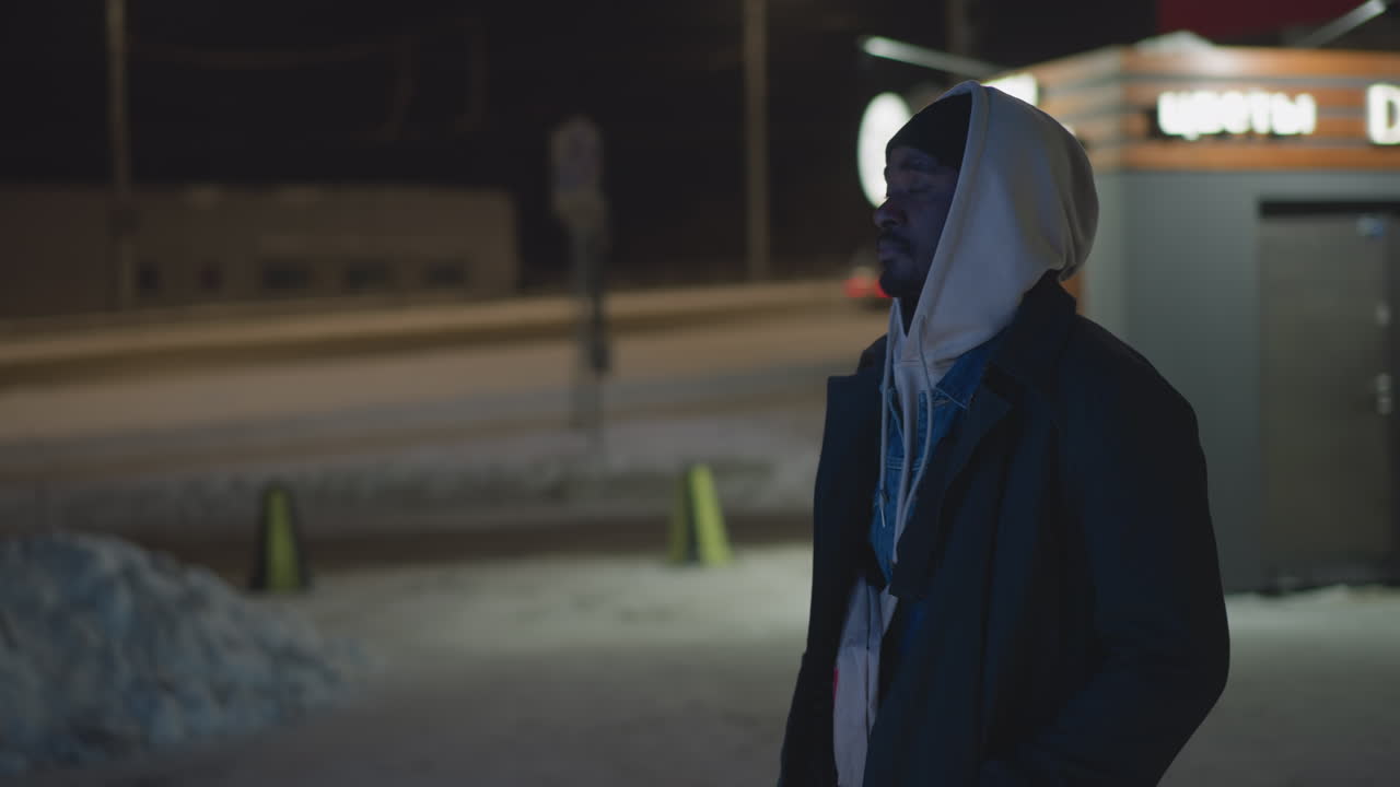 Man standing near glowing Christmas lights in urban night scene. Shop with illuminated signage in background, festive holiday atmosphere, winter setting with street lights and snow