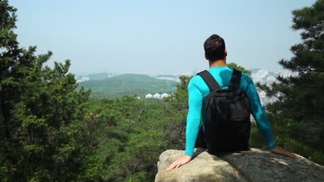 hiker man with backpack sitting on edge of sheer cliff overlooking city panorama