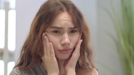 Unhappy-young-woman-looking-at-face-and-touching-skin-front-bathroom-mirror