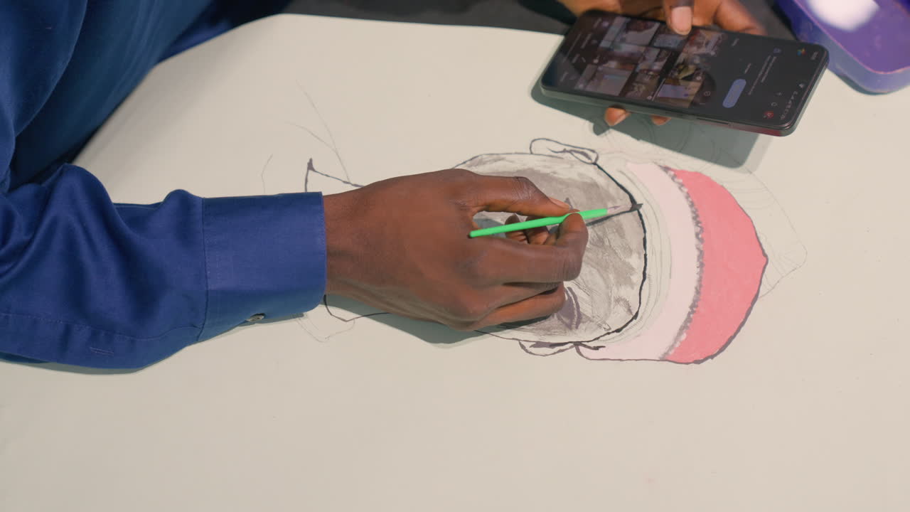 Black artist drawing portrait on paper, closeup of darkskinned hand in blue sleeve using green pen, smartphone reference showing image, visible pink cap in sketch, soft shading and detailed line work