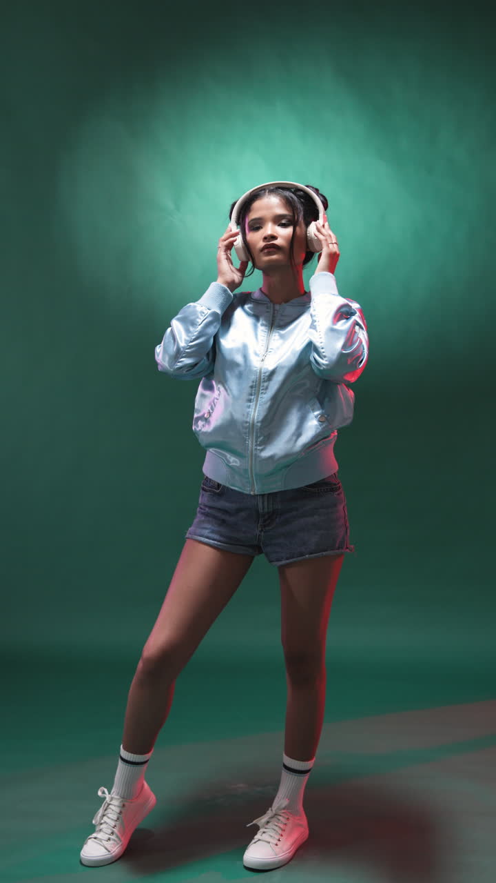 Vertical video: Young woman in headphones dancing in studio wearing denim shorts and jacket