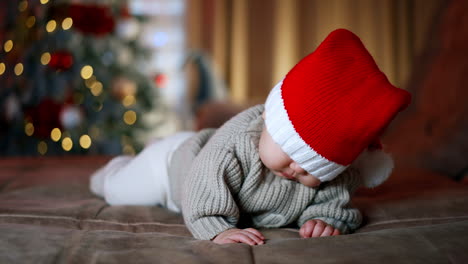 red santa cap covered child's eyes. baby moves to the sides until mom fixes the cap. blurred christmas tree at backdrop.