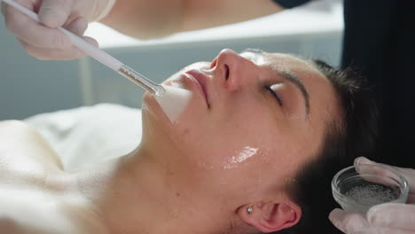 skin care professional in gloves uses brush to apply clear gel mask on closed eyes cheeks and forehead of female model lying relaxed on treatment bed under soft clinical light for deep hydration