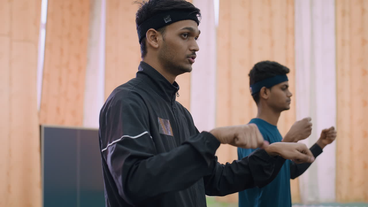 Side view of two tennis players in sportswear warming up with neck flexibility exercise near tennis table, focusing on posture and body readiness before match inside indoor training hall