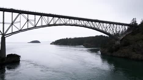 Dark-gray-water,-passing-slowly-under-the-Deception-Pass-Bridge,-Washington-State,-aerial