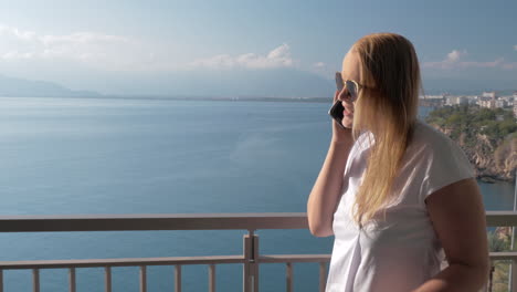 a fair-haired woman talking to a phone on a hotel balcony near the sea landscape