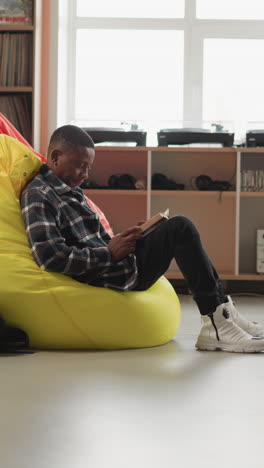 black man enjoys book sitting on bean bag. male reader comfortably sits in cushioned chair in college library lounge engrossed in book. cozy place for education