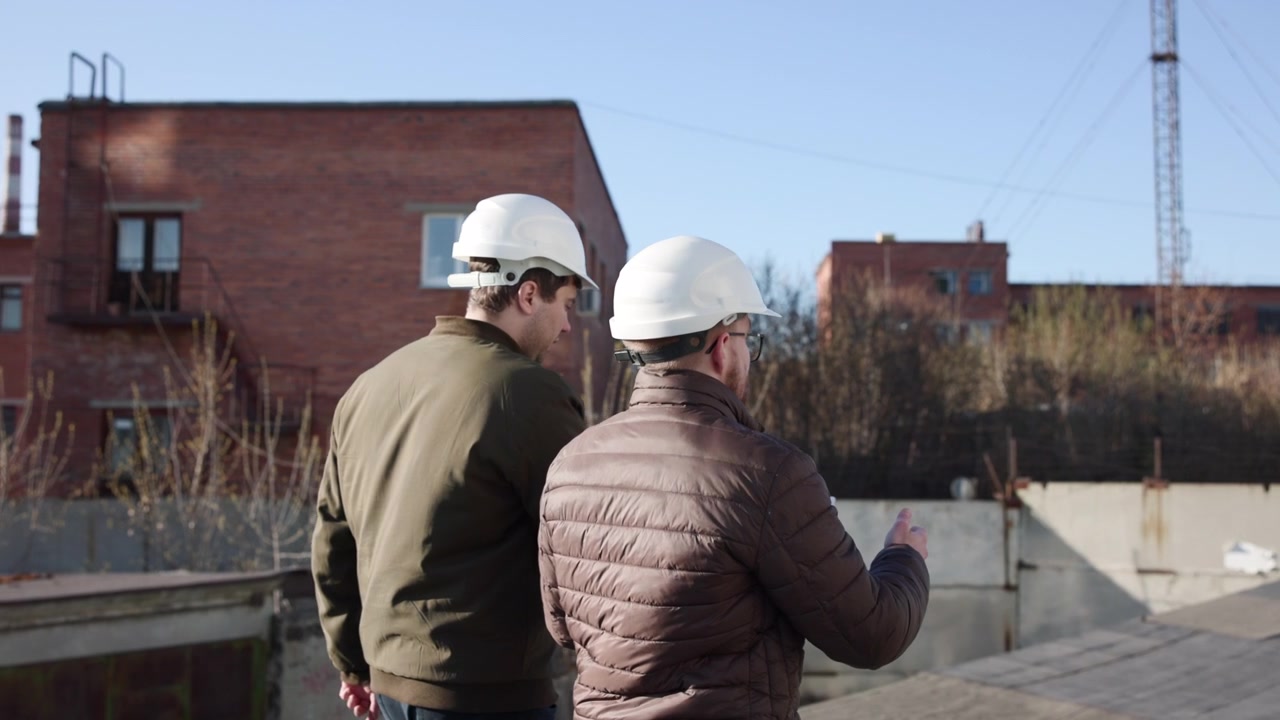 Two Inspectors Walking And Pointing Rooftop, Wearing White Helmets And Casual Jackets, Back View Against Brick. Media