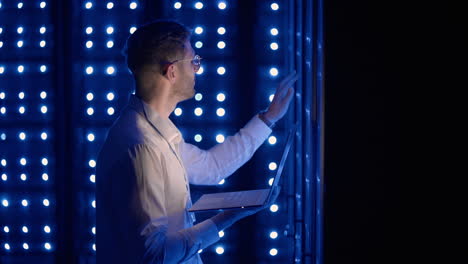 male network engineer doing a system check standing in the server room with his laptop. at data center men server specialists inspecting working system and hardware of rack server computer cabinets