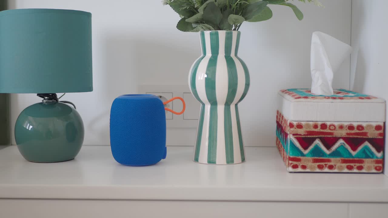 una toma cercana de un altavoz azul, un jarrón verde, una lámpara blanca y una caja de pañuelos en una mesa blanca