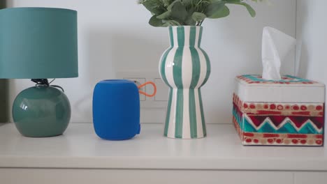 close up shot of a blue speaker, a green vase, a white lamp and a tissue box on a white table