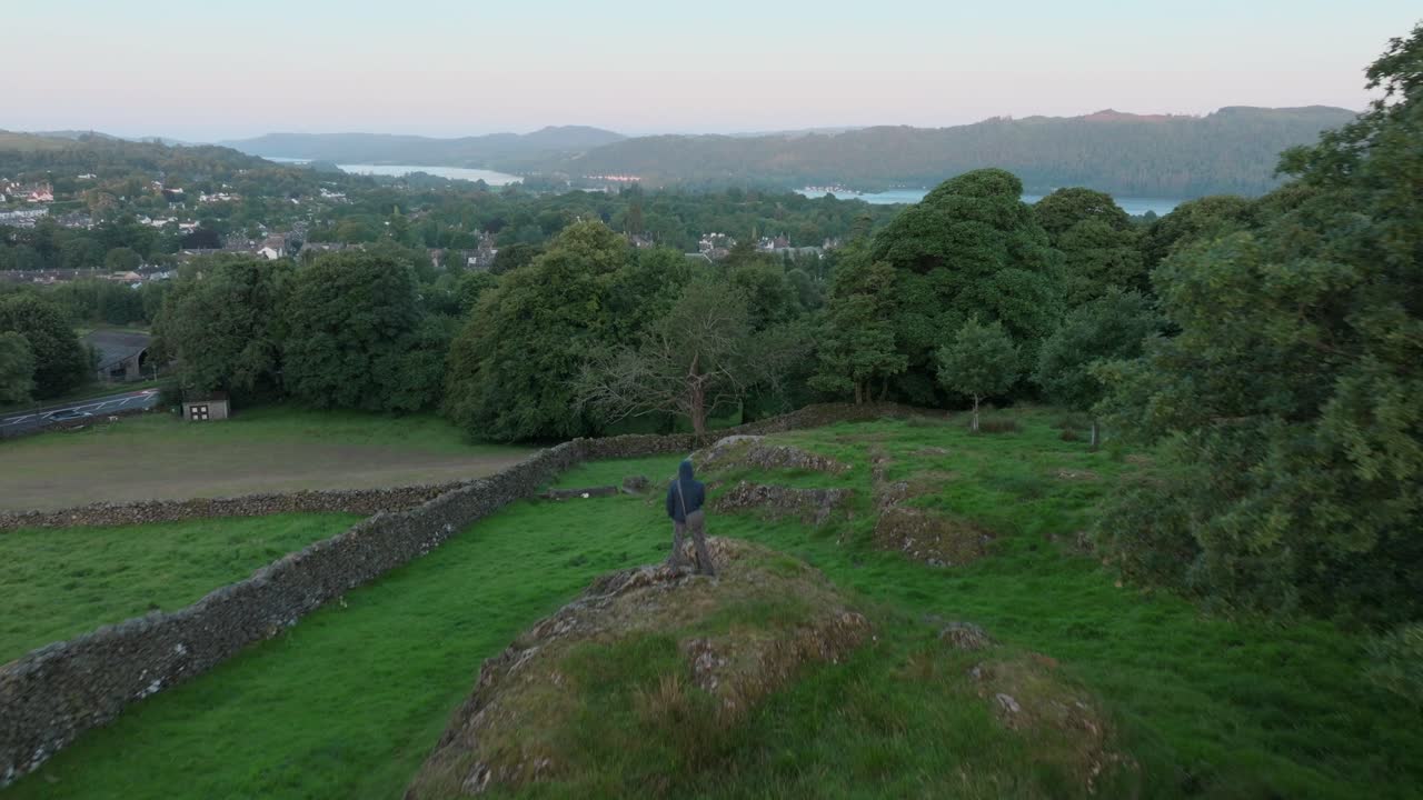 Lone person standing on rocky outcrop with camera flyover towards small woodland and distant lake. Summer. Windermere, Cumbria, UK