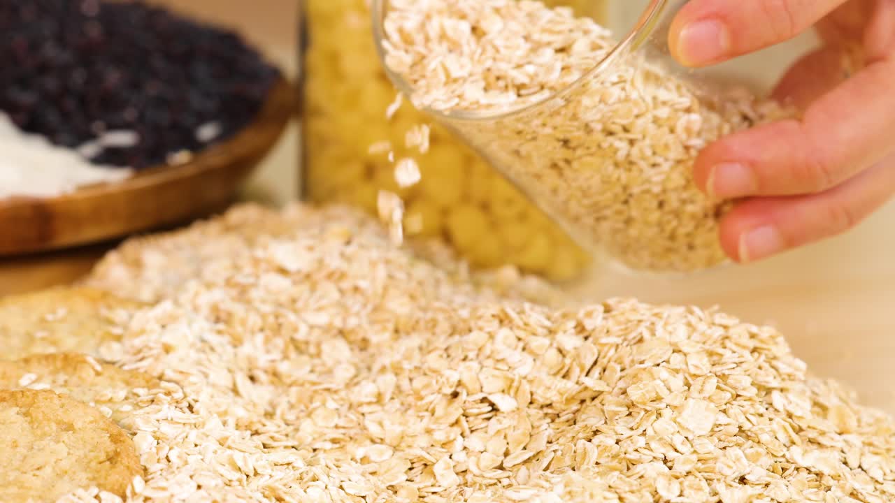 Hands pour rolled oats into a glass container on a kitchen counter. Bright lighting highlights the grains and surrounding ingredients