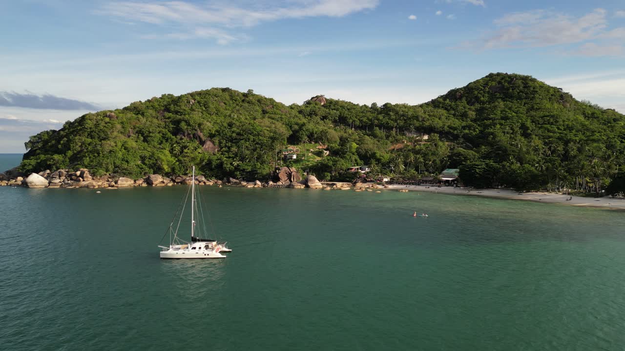 Imagem com zoom lento dos grupos de ilhas Koh Samui durante o dia, com um pequeno iate atracado perto da praia rochosa em águas calmas.
