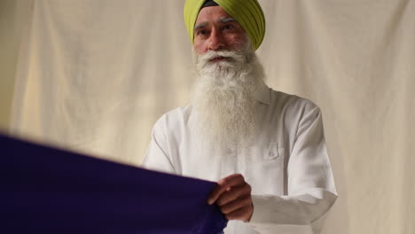 une photo de studio d'un homme sikh senior pliant un tissu pour un turban sur un fond plat 4