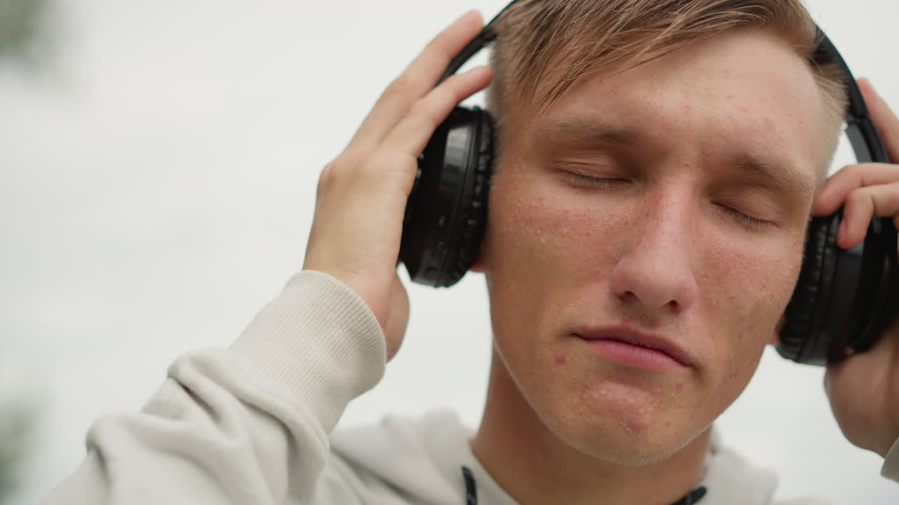 Hombre caucásico en el exterior con auriculares, ajustando los cascos, cerrando los ojos, sintiendo gotas de lluvia en la cara bajo un cielo nublado, sudadera con capucha húmeda por la llovizna, sutil sonrisa de alivio, absorto en la música, expresivo