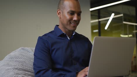 Smiling-mixed-race-businessman-sitting-at-desk-using-laptop-in-office