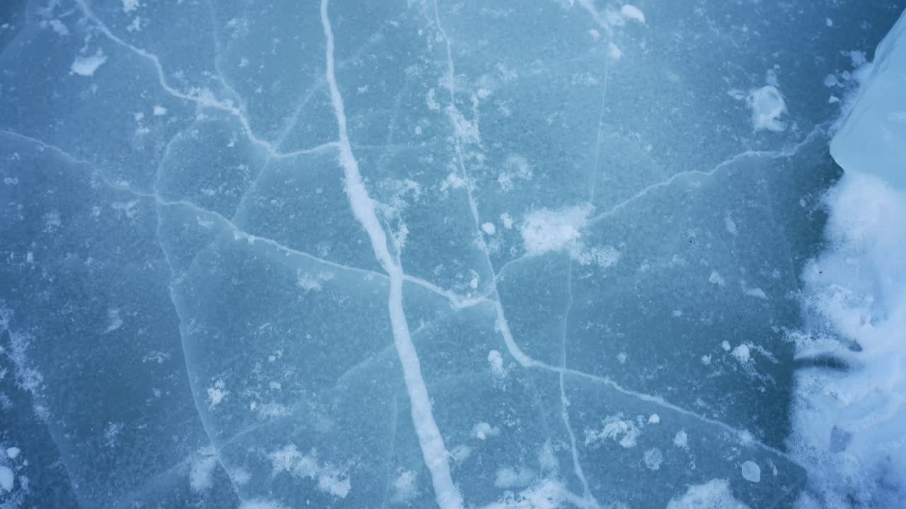Primo piano di ghiaccio blu congelato con lunghe crepe bianche e chiazze di neve sparse. Sfondo astratto con texture invernale che suggerisce il pericolo di ghiaccio sottile e un'atmosfera da clima freddo.