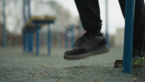 close-up of a person's legs jogging near a blue iron bar and green grass, a bench is visible behind, with a blurred view of a painted yellow house in the distance