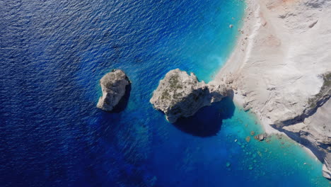 Aéreo:-Toma-De-Arriba-Hacia-Abajo-De-Rocas-De-Piedra-Caliza-De-Myzithres,-Playa-Blanca-Y-Mar-Azul-Bajo-Los-Acantilados-De-La-Isla-De-Zakynthos,-Grecia