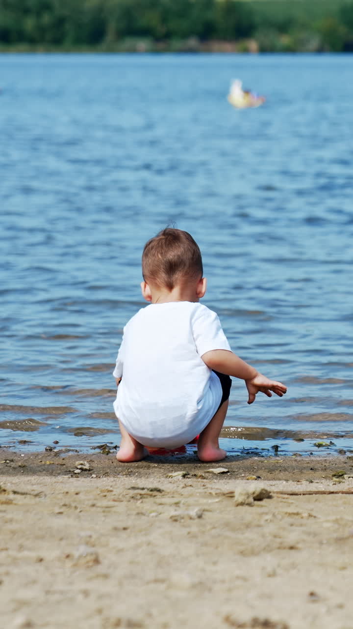 Lovely kid in white T-shirt comes up to a river. Toddler is filling his watering can with water. Vertical video
