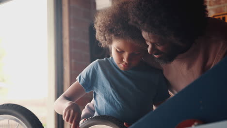 Primer-Plano-De-Un-Niño-Preadolescente-Concentrándose-Mientras-Fija-El-Volante-A-Su-Kart-De-Carreras-En-El-Garaje-Con-Su-Padre-Parado-Detrás-De-él-Ayudando,-Enfoque-Selectivo