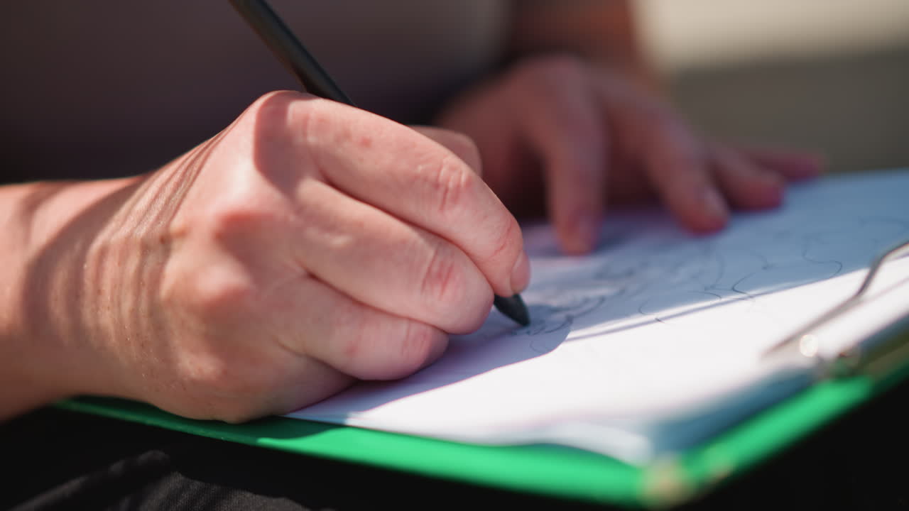 Close up of woman sketching with pencil on paper outdoors, sunlight casting soft hair shadow on hand and page, capturing artistic focus