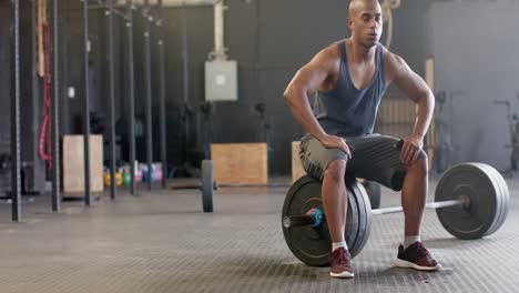 tired biracial man sitting on barbell resting after weight lifting at gym, slow motion, copy space