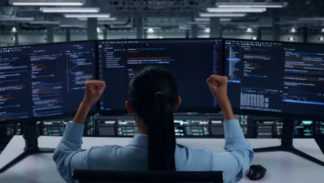vista trasera de una desarrolladora asiática feliz celebrando y bailando mientras escribe código con múltiples pantallas de computadora en el centro de datos
