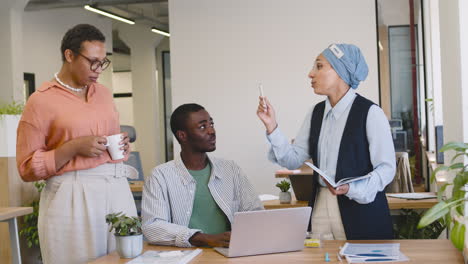 Junger-Arbeiter,-Der-Mit-Einem-Laptop-Arbeitet,-Der-An-Seinem-Schreibtisch-Sitzt,-Während-Eine-Muslimische-Geschäftsfrau-Und-Eine-Geschäftsfrau-Mit-Ihm-Im-Stehen-Sprechen-2