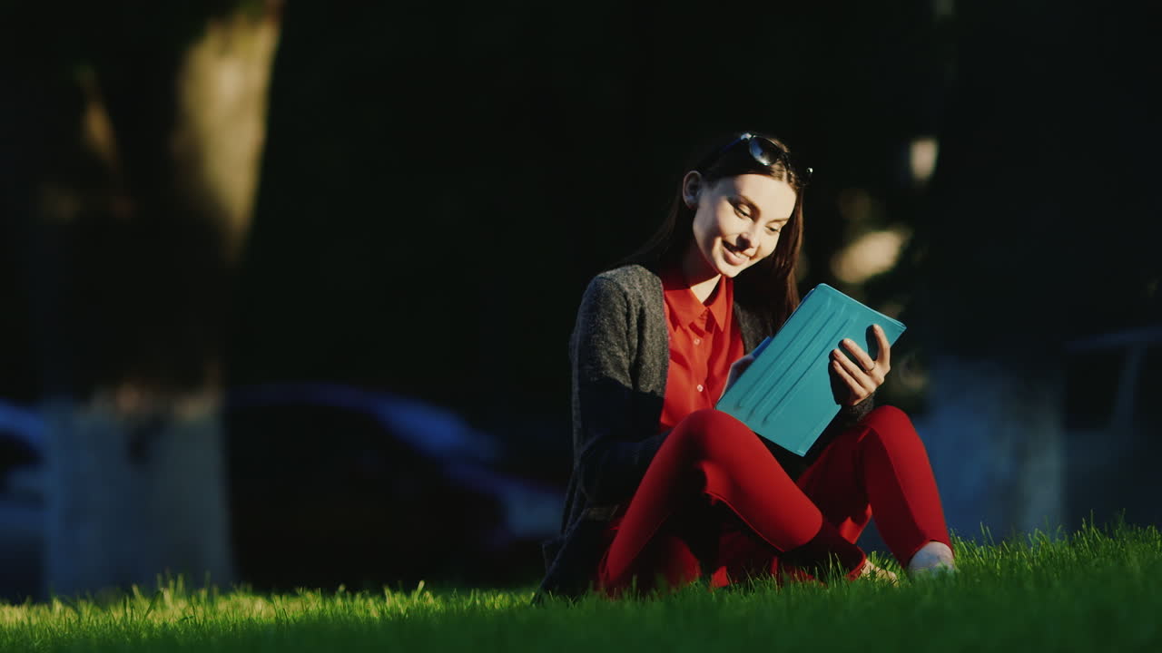mujer joven disfruta de la tableta en el parque sentado en el césped su rostro iluminado por el sol de la tarde hd vi