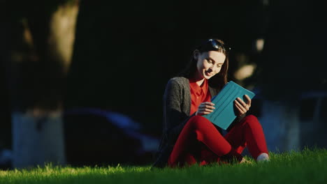 Young-Woman-Enjoys-The-Tablet-In-The-Park-Sitting-On-The-Grass-His-Face-Lit-By-The-Evening-Sun-Hd-Vi