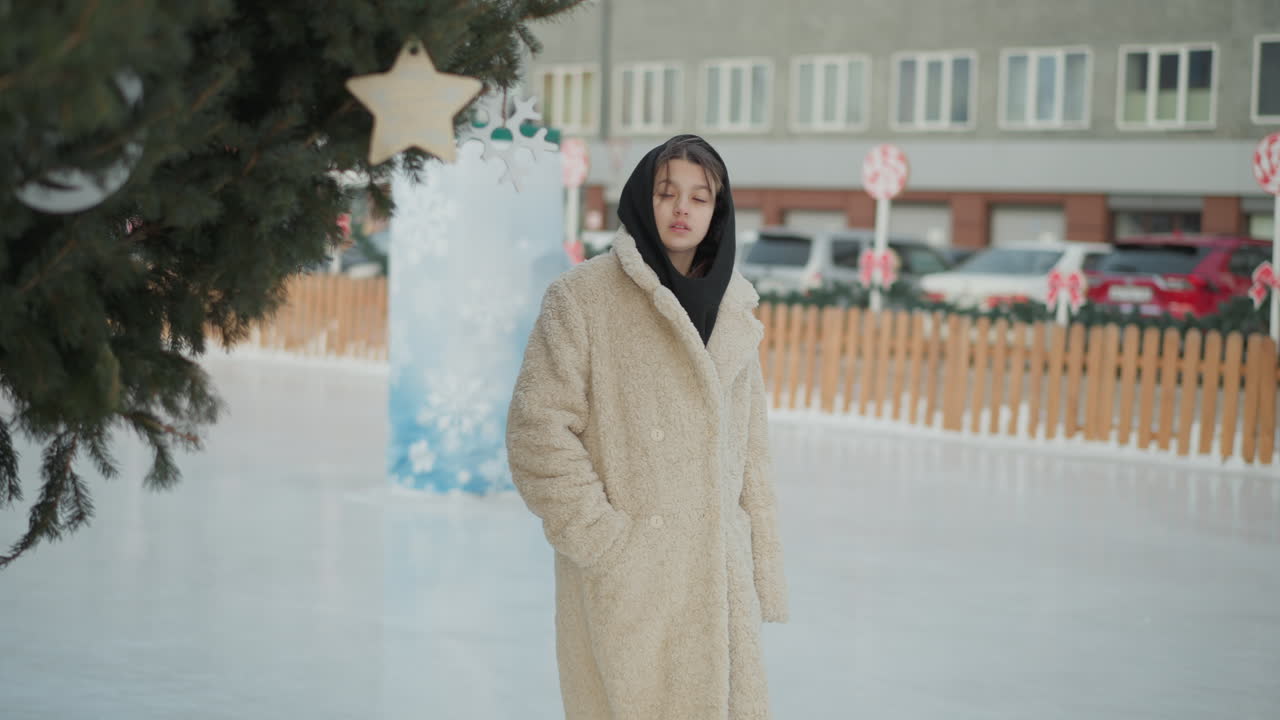 검은 모자를 입은 따뜻한 베이지 겨울 재을 입은 어린 소녀는 눈 인 길에서 부드럽게 고, 초록색, 눈으로 인 땅, 주차된 자동차, 주거 건물을 쳐다보며 평화로운 겨울 도시 풍경을 만니다.