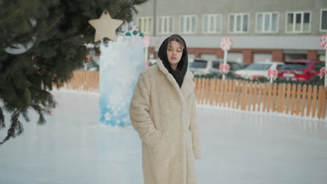 young girl in warm beige winter jacket with black hood walks gently on snowy path, gazing at greenery, snow-covered ground, parked cars, and residential buildings create a peaceful winter cityscape