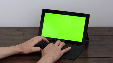 types on a keyboard connected to a tablet with a green screen, stands on a wooden table.