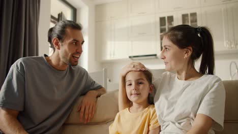 Well,-I'm-a-happy-brunette-guy-in-a-gray-T-shirt-talking-with-his-brunette-girlfriend-and-their-daughter,-a-brunette-girl-in-a-white-T-shirt,-sitting-on-a-brown-sofa-in-a-modern-studio-room