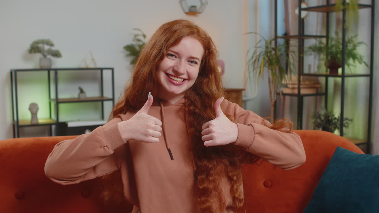 Portrait of smiling caucasian young woman showing double thumbs up sitting on sofa couch at home