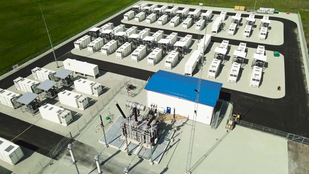 Luftfoto af et moderne BESS, et batterienergilagringssystem til lagring og distribution af vedvarende grøn energi, med rækker af containeriserede batterier og en transformer.