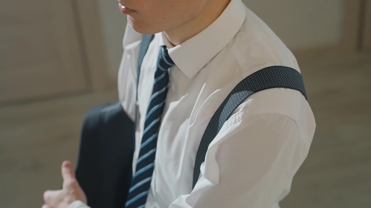 ベストとネクタイを調整するアジア人男性のクローズアップ。明るい室内の床で姿勢を確認しながらジャケットを手に持つ。仕立ての良い生地の質感とストライプのネクタイ。プロフェッショナルな登場に向けた決定的な準備。