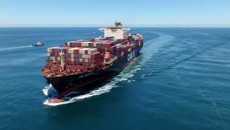 a large container ship navigating through crystal clear waters, surrounded by distant land and tugboats, showcasing its colorful cargo and sleek design