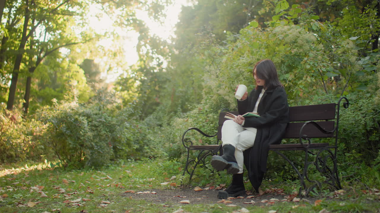 Solitary girl in long jacket, white trousers and black boots seated on bench, reading novel with gentle smile, flipping pages between sips of tea from paper cup, surrounded by autumn greenery