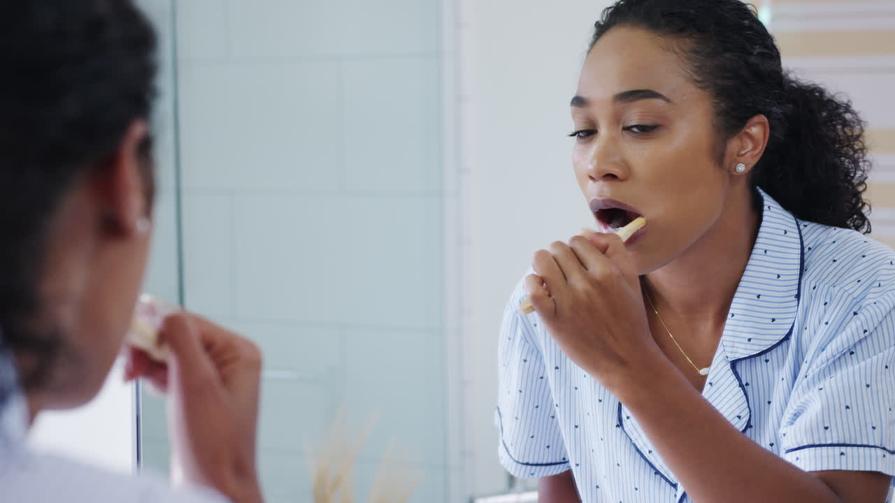 Woman Wearing Pyjamas Standing At Sink Brushing Teeth In Bathroom Reflected In Mirror