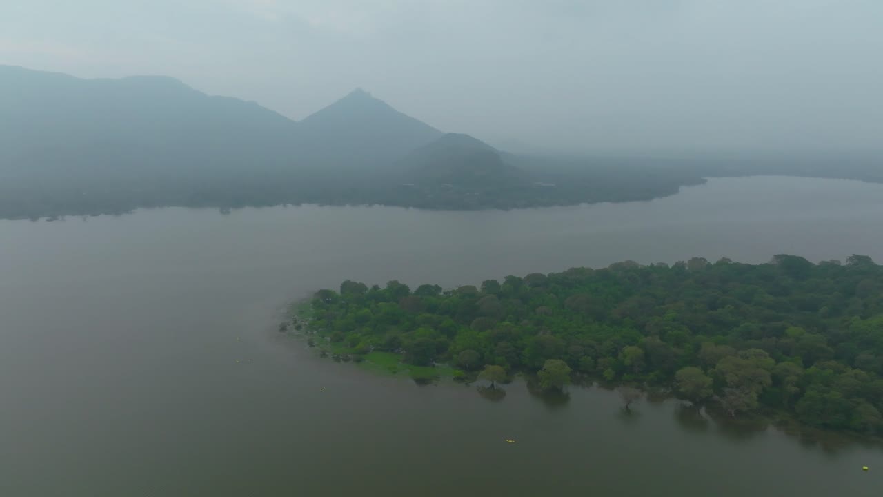 Aerial pan right over lake and tree-covered island, with misty mountains in the background, Sri Lanka