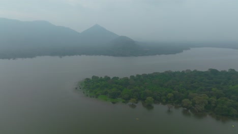 aerial pan right over lake and tree-covered island, with misty mountains in the background, sri lanka